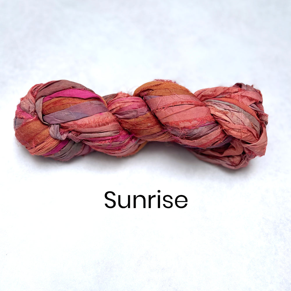 Braided sari silk yarn skein in shades of pink and red and orange on a white background with 'Sunrise' text.