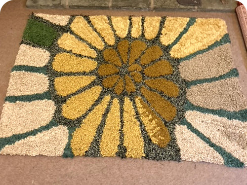 Green and yellow short shaggy rag rugged doormat with sunflower design on a carpeted floor
