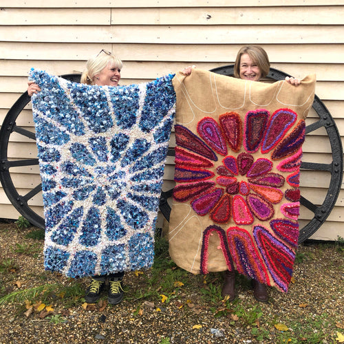 Two people holding up colorful handmade rugs in front of a wooden wall.