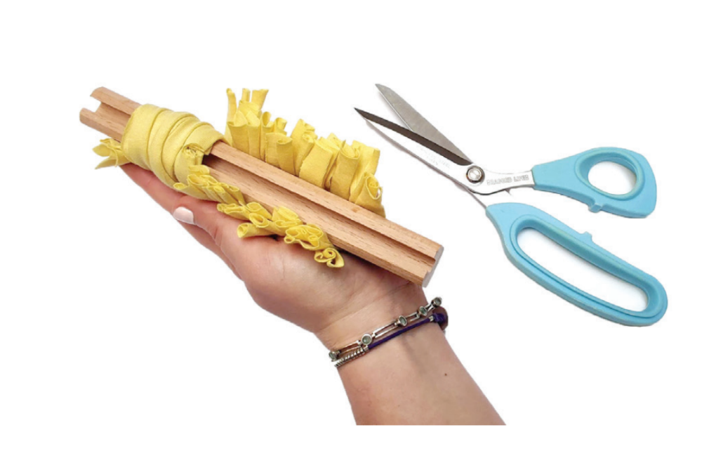 Hand holding Ragged Life rag rug cutting gauge with yellow fabric strips and blue handled fabric scissors