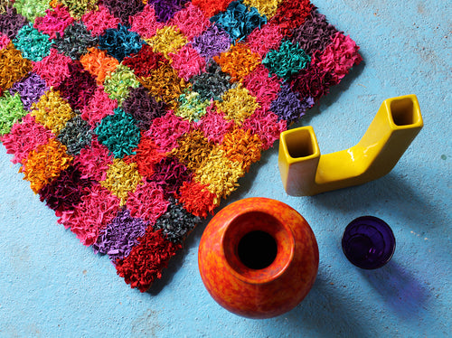 Colorful checked pattern textured rag rug with a yellow vase, red and purple vases on a blue surface
