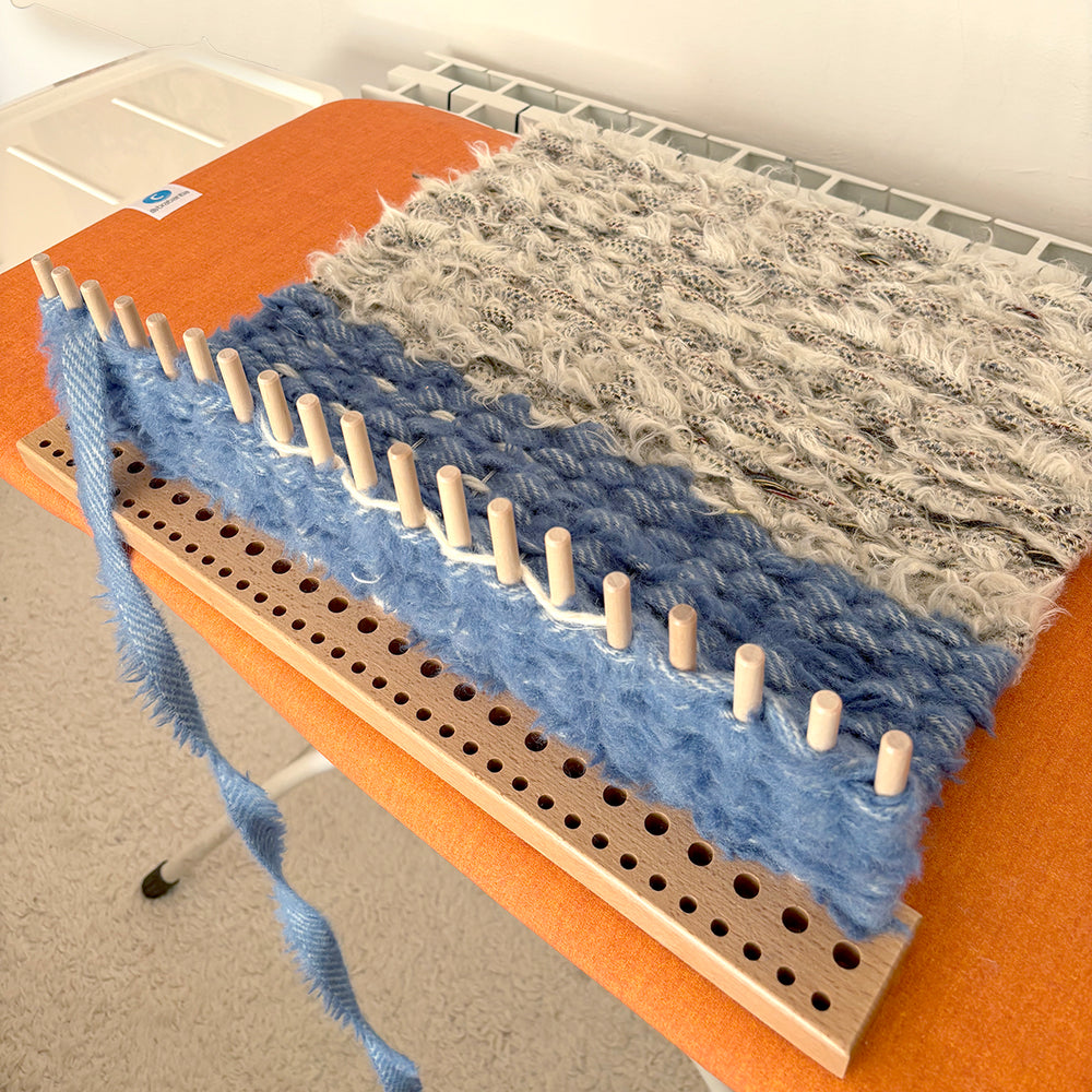 Work in progress peg loom weaving with blue and white woollen blanket selvedge strips on an orange ironing board - great beginner project